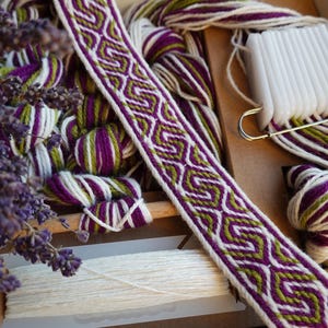 May include: A craft kit featuring purple, green, and white yarn, a woven band, and a white plastic tool. The kit includes dried lavender. The woven band displays a geometric pattern in purple, green, and white.