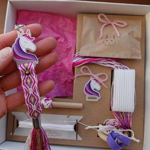 May include: A handmade friendship bracelet kit featuring a unicorn charm. The bracelet has a woven design in shades of pink, purple, and gold. The kit includes a bracelet, additional charms, string, and a wooden tool, all packaged in a box.
