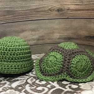 May include: Two crocheted hats in green and brown. One hat is a simple beanie style, while the other is shaped like a turtle shell.
