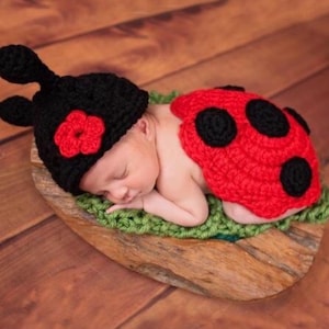 May include: A newborn baby wearing a handmade crochet ladybug costume, including a black hat with red flower and antennae, and a red shell with black spots. The baby is lying on a green crocheted surface inside a wooden bowl.