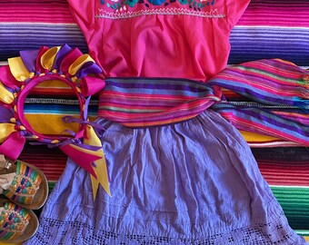 Può includere: Una gonna viola con un bordo di pizzo bianco, una blusa rosa ricamata con maniche corte, una fascia a righe colorate e una fascia per capelli gialla e viola. L'abito è disposto su una coperta a righe colorate.