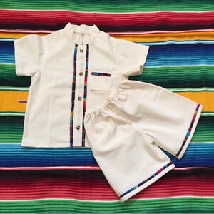 May include: A white two-piece outfit for a child, consisting of a short-sleeved shirt with a mandarin collar and a pair of shorts. The shirt and shorts have colorful trim along the edges.