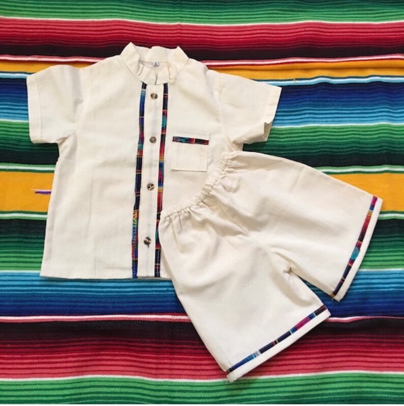 May include: A white two-piece outfit for a child, consisting of a short-sleeved shirt with a mandarin collar and a pair of shorts. The shirt and shorts have colorful trim along the edges.