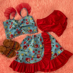 May include: A blue and red skirt with a mermaid print and red ruffle trim. The skirt is paired with a matching blue crop top with red straps and a red ruffle trim. The outfit is completed with a pair of red bloomers and brown sandals.