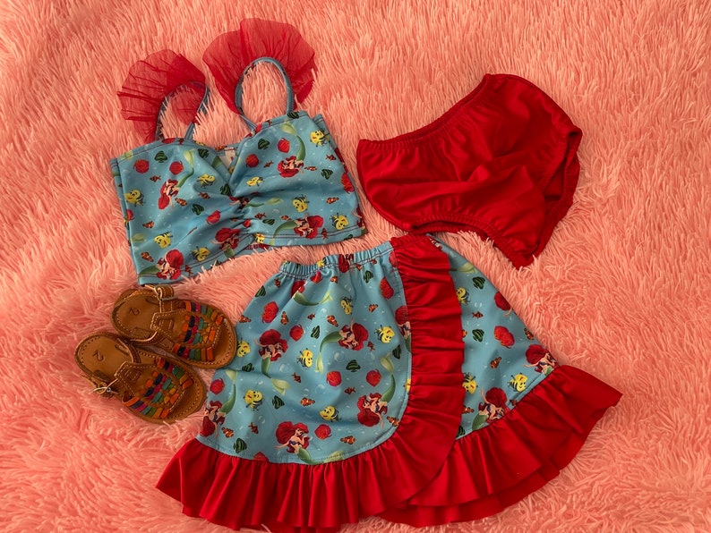May include: A blue and red skirt with a mermaid print and red ruffle trim. The skirt is paired with a matching blue crop top with red straps and a red ruffle trim. The outfit is completed with a pair of red bloomers and brown sandals.