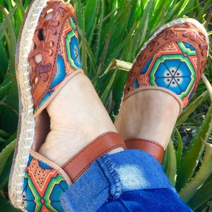 May include: Handmade leather espadrille sandals with colourful geometric embroidery. The sandals feature a brown leather upper with cut-out details, a woven jute sole, and a strap. The embroidery includes blue, green, orange, and yellow patterns.