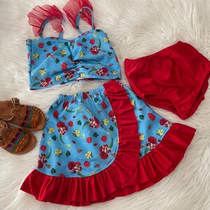 May include: A blue and red two-piece swimsuit with a mermaid print. The top has a ruffled neckline and the skirt has a ruffled hem. The swimsuit is paired with red diaper-style bottoms.