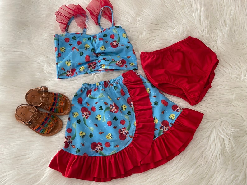 May include: A blue and red two-piece swimsuit with a mermaid print. The top has a ruffled neckline and the skirt has a ruffled hem. The swimsuit is paired with red diaper-style bottoms.