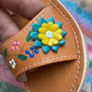Pu&ograve; includere: Sandali in pelle color cuoio con un design floreale. I fiori sono dipinti in tonalit&agrave; di rosa, blu, verde, giallo e bianco.