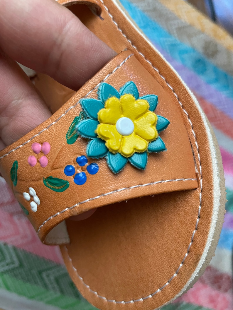 Pu&ograve; includere: Sandali in pelle color cuoio con un design floreale. I fiori sono dipinti in tonalit&agrave; di rosa, blu, verde, giallo e bianco.