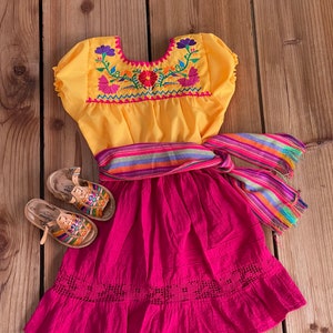 May include: A yellow embroidered dress with a pink skirt and a colorful striped sash. The dress has short puffed sleeves and a round neckline. The skirt has a scalloped hem and a lace trim. The dress is paired with a pair of brown leather sandals.