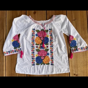 Peut inclure: Chemise blanche brod&eacute;e avec un motif floral dans des tons de rose, jaune, bleu et vert. La chemise a des manches longues avec des glands roses.