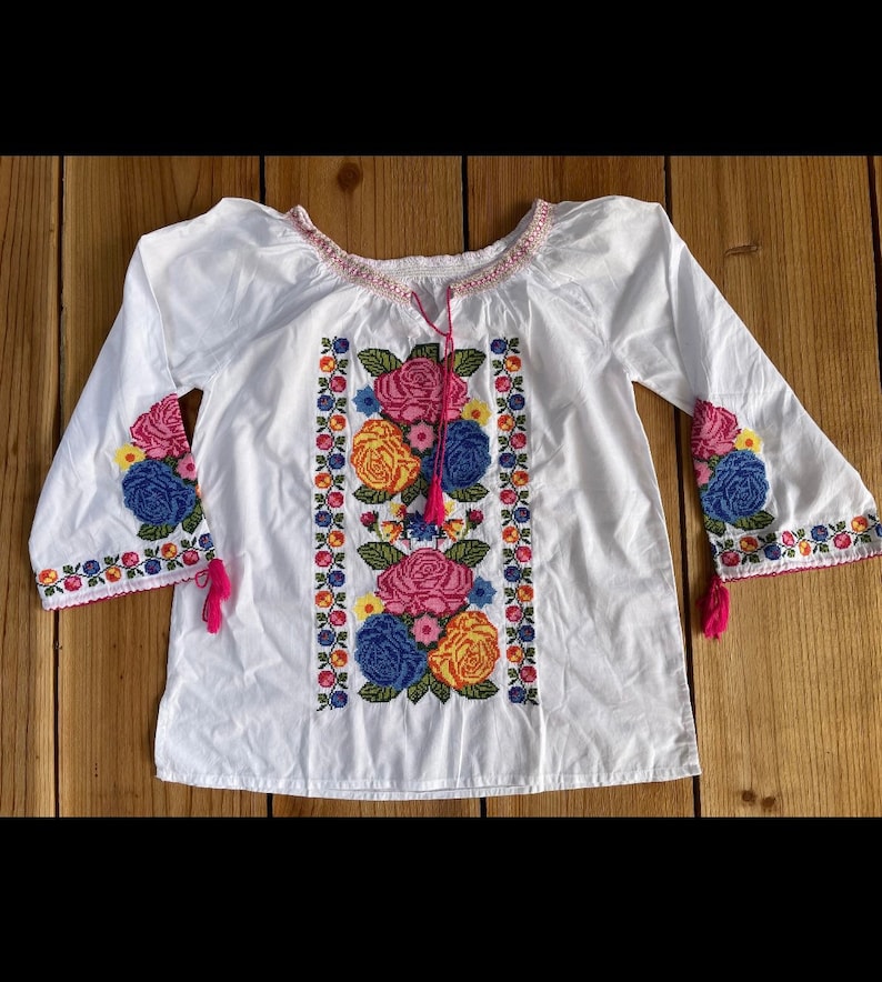 Peut inclure: Chemise blanche brod&eacute;e avec un motif floral dans des tons de rose, jaune, bleu et vert. La chemise a des manches longues avec des glands roses.