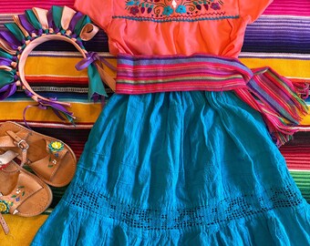 Può includere: Un vestito ricamato turchese e corallo con una cintura a righe rosa, viola e verde. Il vestito ha una gonna a balze ed è decorato con ricami floreali. Il vestito è steso su una coperta a righe colorate.