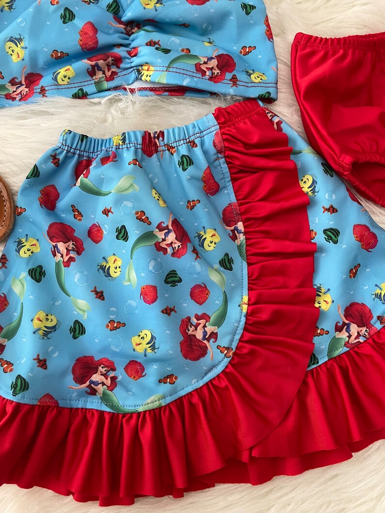 May include: A blue and red two-piece swimsuit with a mermaid and fish print. The top is a bandeau style and the bottom is a skirt with a ruffle trim.