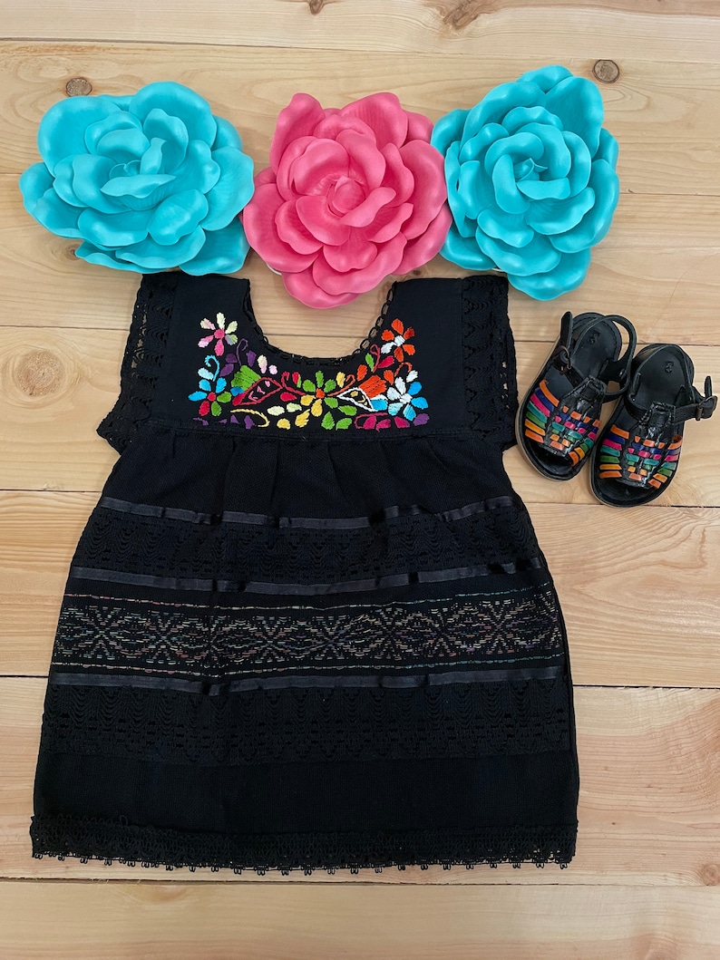 May include: Black embroidered dress with colorful floral and bird designs, featuring a lace trim and multiple layers of black ribbon. The dress is displayed on a wooden surface with three paper flowers.