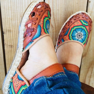May include: A pair of colourful espadrille sandals with a woven jute sole. The sandals feature a brown leather upper with a geometric pattern in blue, green, red, and yellow. The sandals are worn with blue denim jeans.