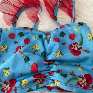 May include: A blue swimsuit top with a red ruffle trim and a mermaid and fish print. The top has a V-neck and adjustable straps.