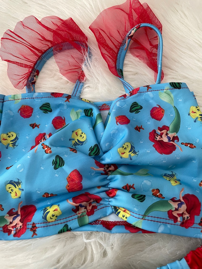 May include: A blue swimsuit top with a red ruffle trim and a mermaid and fish print. The top has a V-neck and adjustable straps.