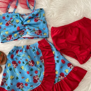 May include: A three-piece swimsuit for girls featuring a red and blue mermaid print. The top has red tulle straps and a ruffled trim. The skirt is a wrap style with a red ruffle trim. The bottoms are solid red.