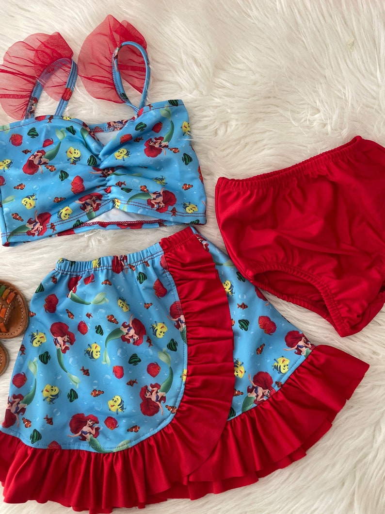 May include: A three-piece swimsuit for girls featuring a red and blue mermaid print. The top has red tulle straps and a ruffled trim. The skirt is a wrap style with a red ruffle trim. The bottoms are solid red.