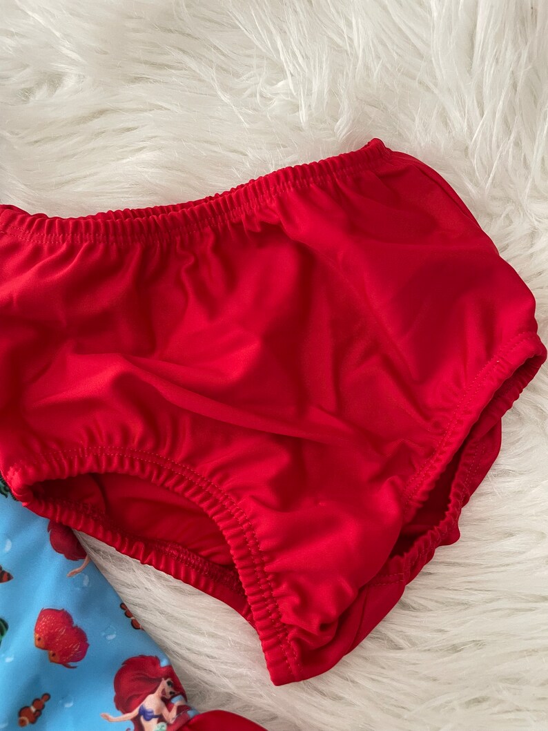 May include: Red cotton diaper with elastic waistband. The diaper is made of a soft, stretchy fabric.