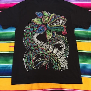 May include: Black t-shirt featuring a colorful illustration of a serpent-like creature. The design incorporates shades of green, blue, pink, and yellow. The graphic is detailed with intricate patterns and a stylized appearance. The shirt is laid flat on a striped textile.