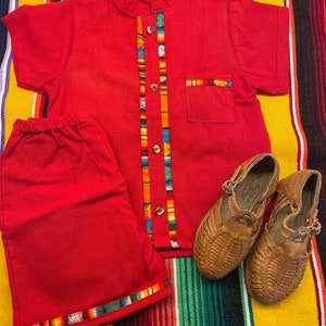 May include: Red short-sleeved shirt with a colorful trim and matching red shorts. The shirt has a mandarin collar and button closure. The shorts have a colorful trim around the bottom hem. Two pairs of brown leather sandals are also pictured.