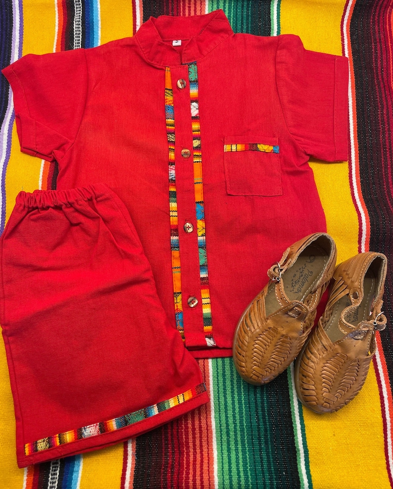 May include: Red short-sleeved shirt with a colorful trim and matching red shorts. The shirt has a mandarin collar and button closure. The shorts have a colorful trim around the bottom hem. Two pairs of brown leather sandals are also pictured.