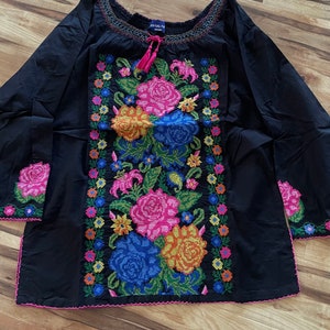 Peut inclure: Chemise noire &agrave; manches longues avec un motif de broderie florale. La broderie pr&eacute;sente des fleurs color&eacute;es, notamment roses, jaunes, oranges, bleues et vertes. La chemise a un col fronc&eacute; et un lien au cou.