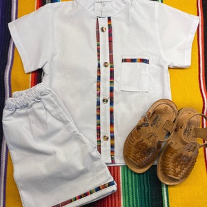 May include: White short-sleeved shirt with a colorful trim down the front and a matching pair of shorts. The shirt has a mandarin collar and a small pocket on the left chest. The shorts have a colorful trim around the bottom. The outfit is laid out on a colorful striped blanket.