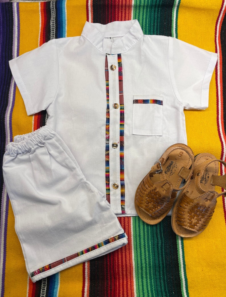 May include: White short-sleeved shirt with a colorful trim down the front and a matching pair of shorts. The shirt has a mandarin collar and a small pocket on the left chest. The shorts have a colorful trim around the bottom. The outfit is laid out on a colorful striped blanket.