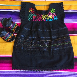 May include: A black embroidered dress with colorful floral details and lace trim. The dress is on a colorful striped blanket.