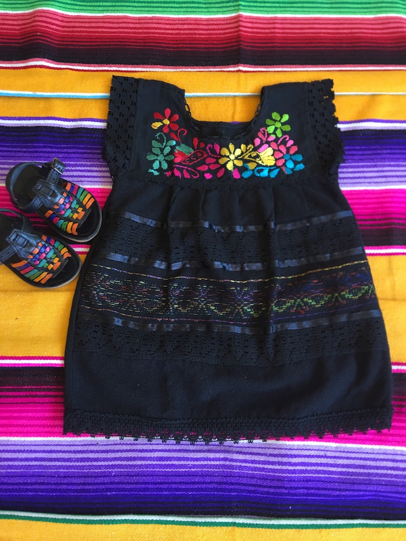 May include: A black embroidered dress with colorful floral details and lace trim. The dress is on a colorful striped blanket.