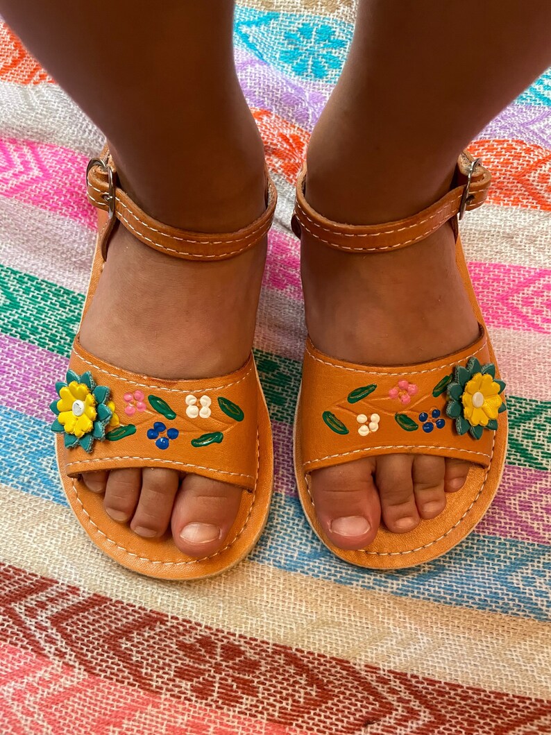 Pu&ograve; includere: Un paio di sandali in pelle marrone con ricami floreali colorati. I sandali hanno un cinturino che va intorno alla caviglia e una chiusura a fibbia.