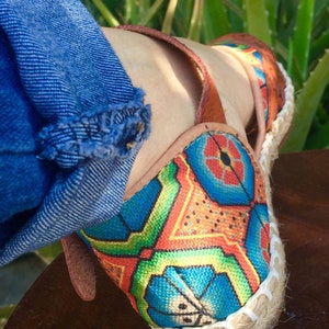 May include: Close-up of a colourful espadrille shoe with a geometric pattern in blue, orange, green, and red. The shoe has a woven jute sole and brown leather straps. Denim jeans are visible in the background.