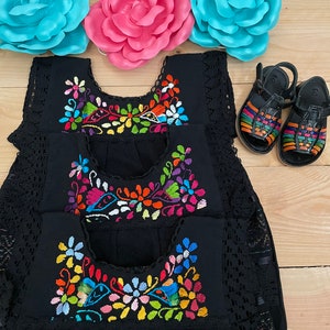 May include: Three black sleeveless dresses with colorful embroidered floral designs. The dresses have a lace trim and are layered on top of each other. Two pairs of black sandals with colorful straps are in the background.