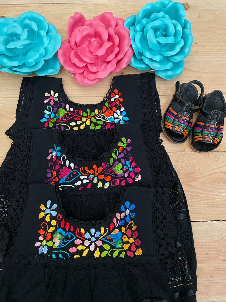 May include: Three black sleeveless dresses with colorful embroidered floral designs. The dresses have a lace trim and are layered on top of each other. Two pairs of black sandals with colorful straps are in the background.