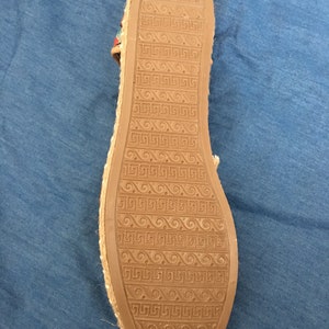 May include: Underside of a tan espadrille sandal with a brown rubber sole. The sole has a repeating geometric pattern. The sandal's edge is made of woven material, and the strap has colourful accents.