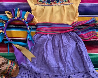 Può includere: Un completo abito tradizionale messicano, con un top giallo ricamato, una gonna viola e una fascia a righe colorata. Una fascia abbinata con nastri e sandali in pelle intrecciata completano l'ensemble. L'outfit è esposto su una coperta a righe.