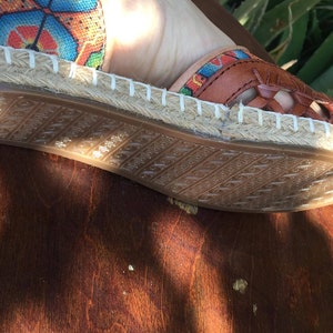 May include: Espadrille sandal with a woven leather upper in shades of brown. The sandal features a colourful textile accent and a jute rope sole. The rubber sole has a textured pattern. The sandal is designed for warm weather.