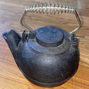 May include: Black cast iron kettle with a gold metal handle and a spring-loaded lid. The kettle has a rounded body and a spout on the side.