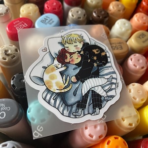 May include: A die-cut sticker featuring a cartoon illustration of two people cuddling in bed with a calico cat and a ginger tabby cat. The characters have rosy cheeks and are wearing pajamas, resting on a blue and white striped blanket with a white pillow.