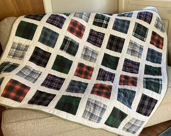 May include: A handmade quilt with a patchwork design, featuring squares of plaid fabric in various colors like red, green, blue, and gray. The quilt is draped over a beige armchair, showcasing its cozy and decorative appeal.