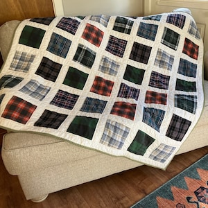 May include: A handmade quilt with a patchwork design, featuring squares of plaid fabric in various colors like red, green, blue, and gray. The quilt is draped over a beige armchair, showcasing its cozy and decorative appeal.