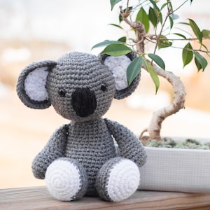 May include: A handmade gray crochet koala bear with white accents on the ears and feet. The koala is sitting in front of a small bonsai tree in a white rectangular pot, placed on a wooden surface.