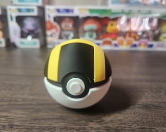 Könnte beinhalten: Ein schwarzer und gelber Pokéball mit einem weißen Knopf in der Mitte. Der Pokéball ist ein beliebtes Spielzeug und Sammlerstück für Fans des Pokémon-Franchise.