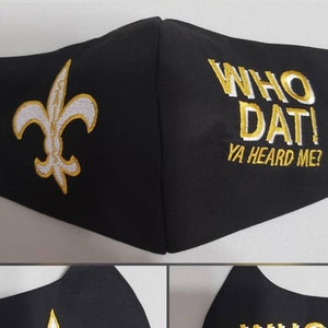 Op de afbeelding: Zwart gezichtsmasker met een witte fleur-de-lis en de tekst "WHO DAT! YA HEARD ME?" in gouden borduurwerk.