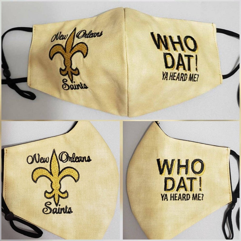 Op de afbeelding: Een beige gezichtsmasker met zwarte bandjes. Het masker is geborduurd met het logo van de New Orleans Saints en de tekst "WHO DAT! YA HEARD ME?" in goud.