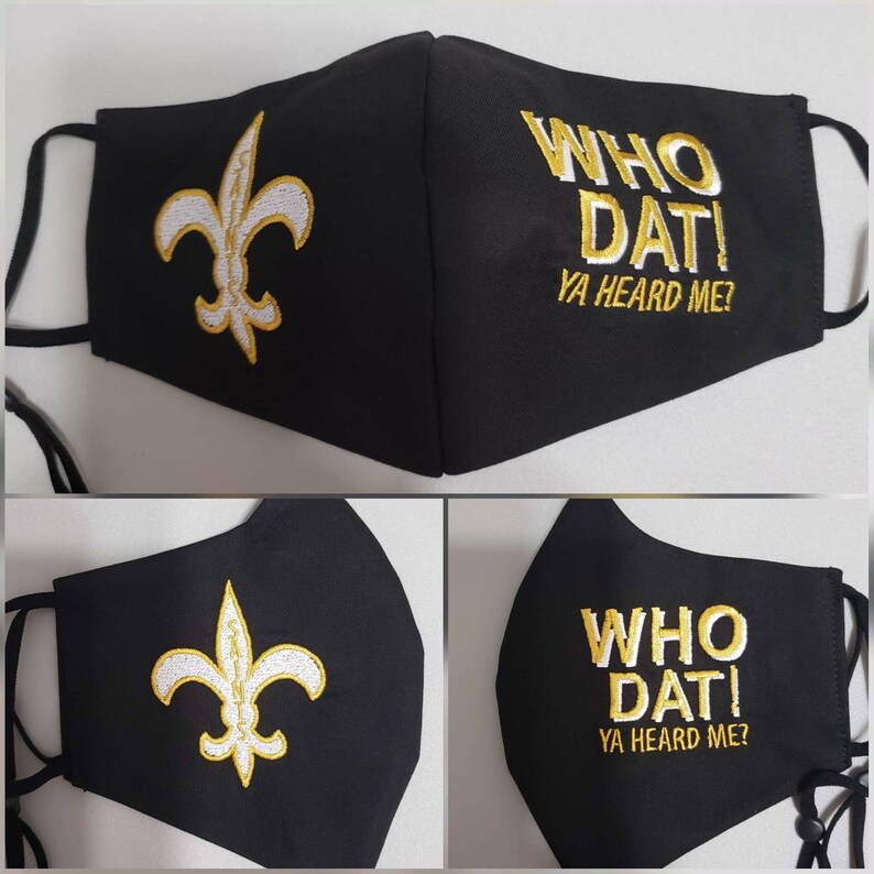 Op de afbeelding: Zwart gezichtsmasker met een witte fleur-de-lis en de tekst "WHO DAT! YA HEARD ME?" in gouden borduurwerk.